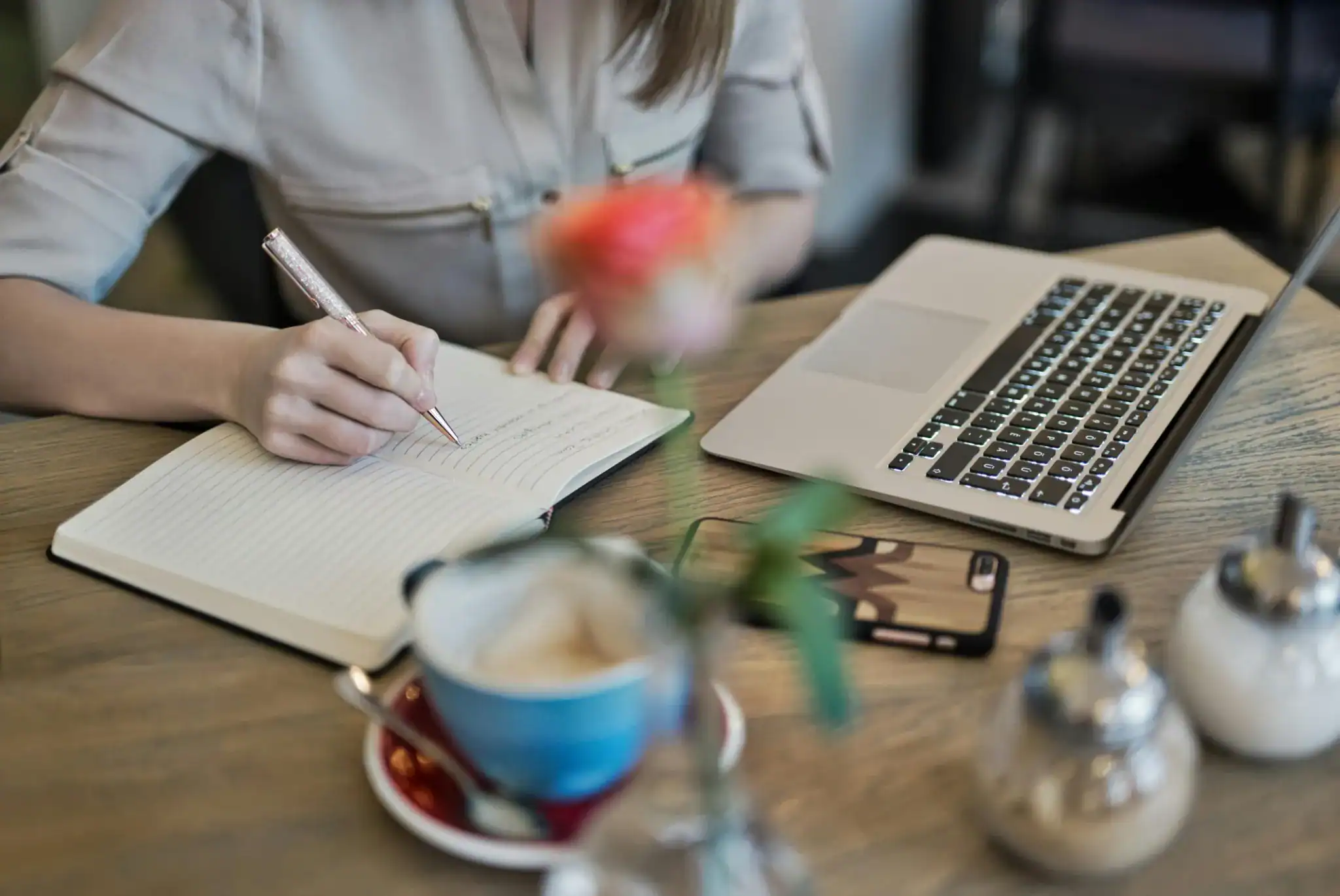 A woman writing in a journal beside a laptop, coffee mug, and flower vase on a wooden desk — symbolizing mindful remote work.