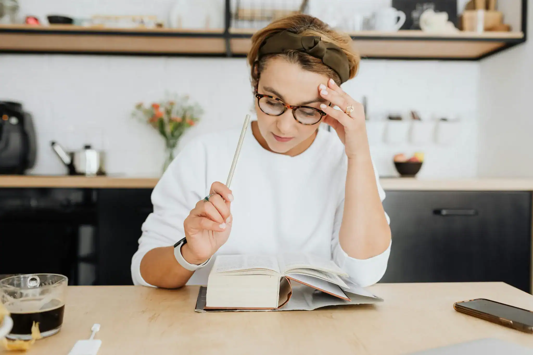 Focused woman reading a book at a cozy kitchen table with a phone placed face down.