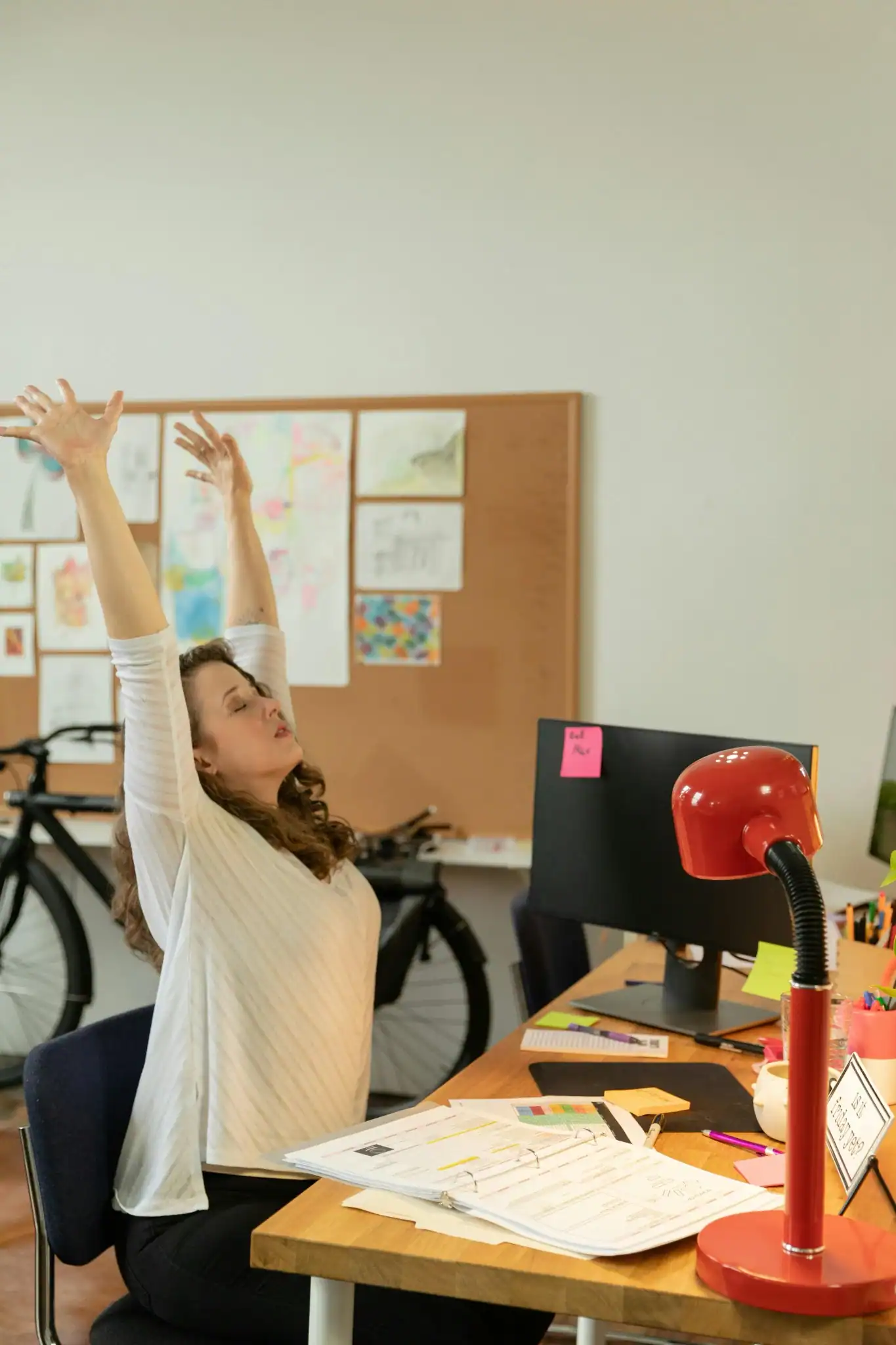 A woman stretching her arms upward while sitting at her desk in a home office, surrounded by papers and a computer—taking a mindful break during remote work.