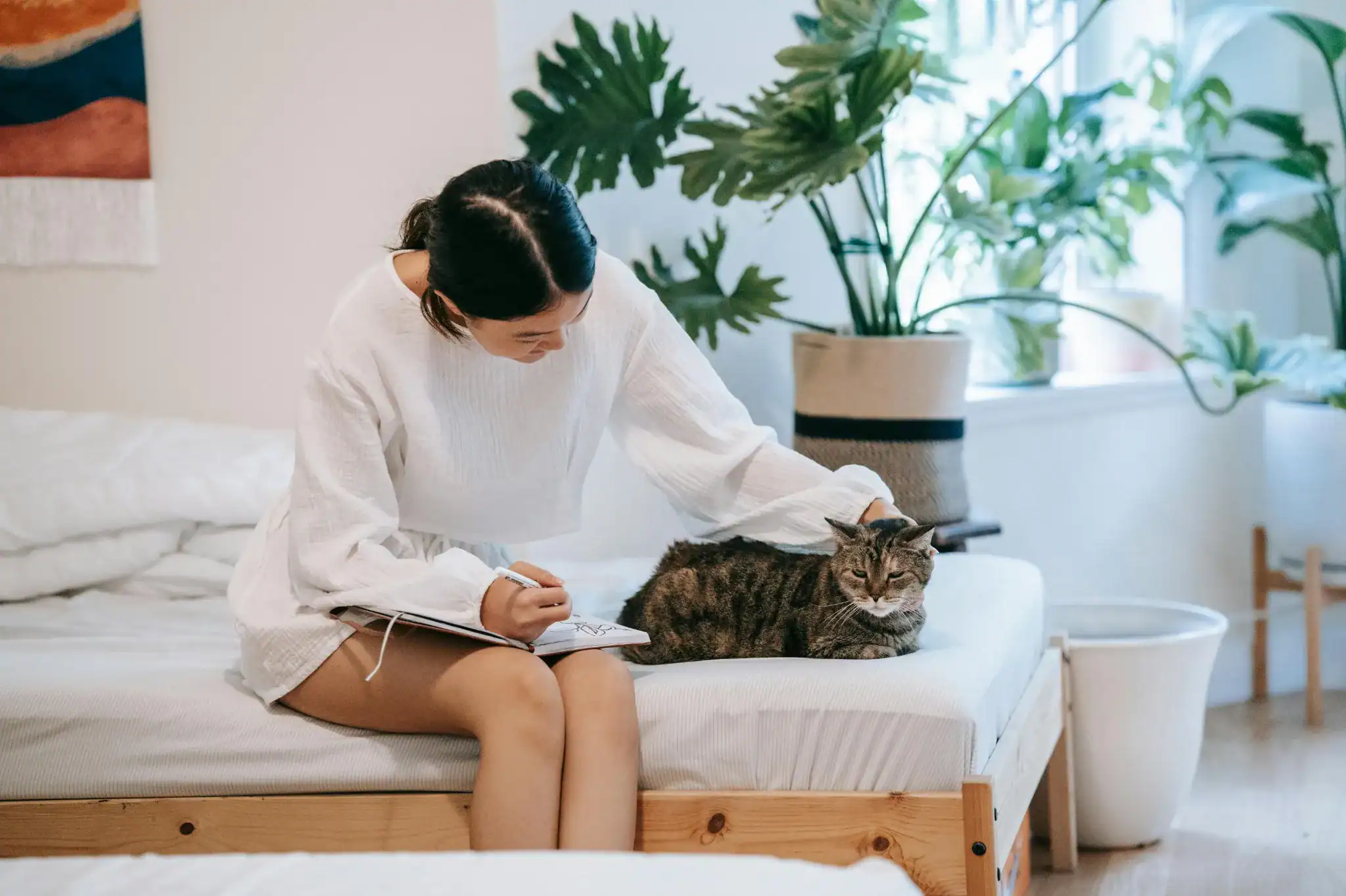 "Young woman journaling on bed while petting a cat, surrounded by indoor plants in a calm, cozy room – symbolizing weekend relaxation and offline detox."